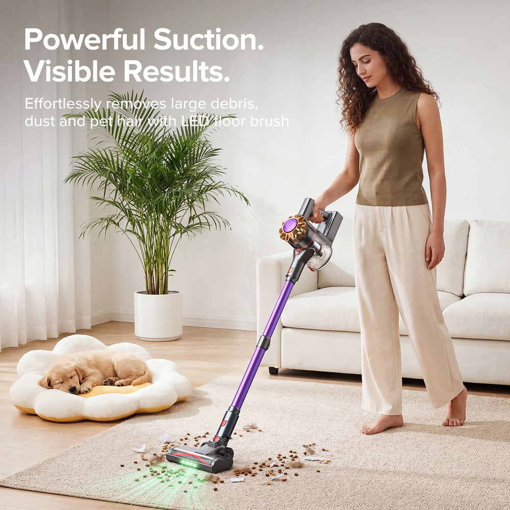 A woman stands barefoot in a bright living room using a purple and gray cordless stick vacuum with a pet hair pickup tool attachment. A small dog sleeps in a cream flower-shaped pet bed on the left, and the vacuum is shown picking up scattered kibble and pet hair from a beige rug.
