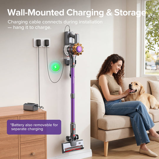 A wall-mounted charging and storage unit holding a purple cordless stick vacuum cleaner, shown in a bright living room with a woman and dog sitting on a beige sofa. White text at the top explains wall-mounted charging and storage, and a purple callout at the bottom mentions removable battery charging.