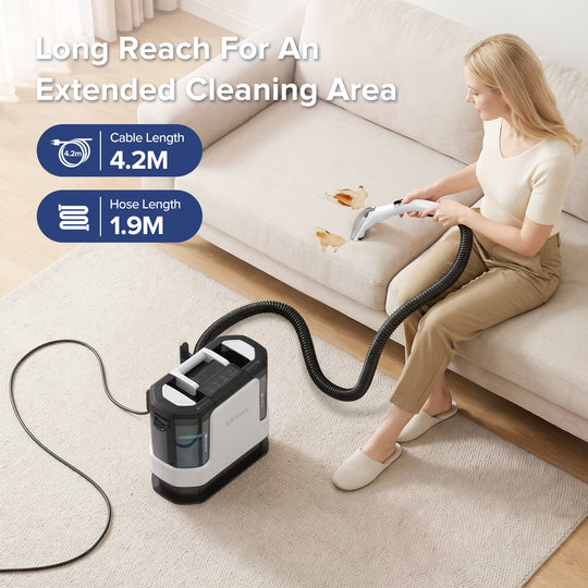 A woman sits on a beige sofa using a cordless floor cleaner with an extended hose and cable on a light rug. The cleaner is black and white with a transparent water tank.